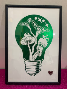 Shiny green foil art print. Hand drawn mushroom elements within a light bulb illustration. A4 sized art print. With Love From Lilibet logo. Foil print in a narrow black frame balanced on a pink glitter table top