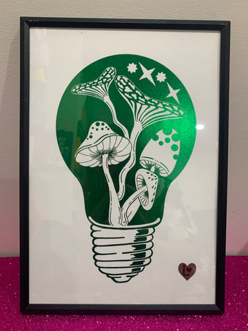 Shiny green foil art print. Hand drawn mushroom elements within a light bulb illustration. A4 sized art print. With Love From Lilibet logo. Foil print in a narrow black frame balanced on a pink glitter table top